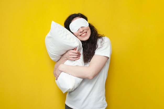 Femme en masque de sommeil étreint un oreiller doux blanc sur fond jaune et dort