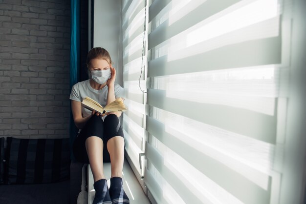 Femme en masques médicaux de protection est assis sur le rebord de la fenêtre et lit un livre