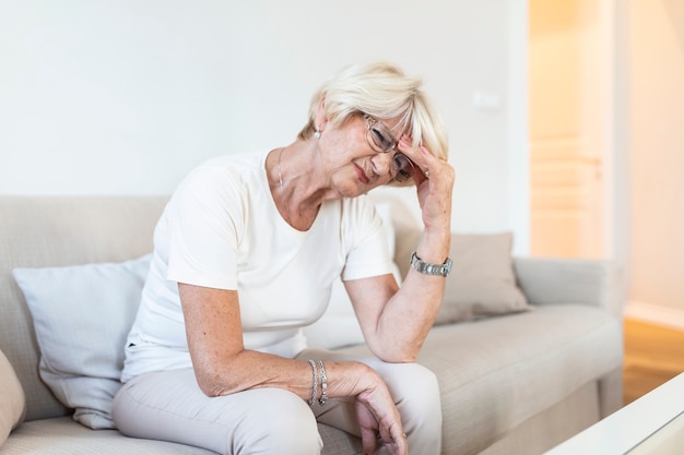 Femme mature assise sur un canapé blanc dans une maison touchant sa tête avec sa main tout en ayant mal à la tête et en se sentant mal