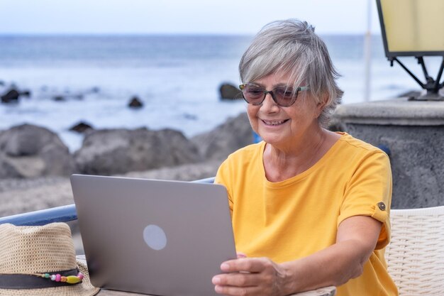 Femme mature assise en plein air en mer sur une table basse à l'aide d'un ordinateur portable. Blogueur, voyageur, tech et social.