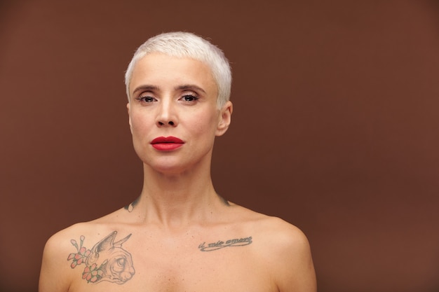 Femme mature aux cheveux blonds courts, tatouages sur le cou et la poitrine et rouge à lèvres rouge sur les lèvres vous regarde