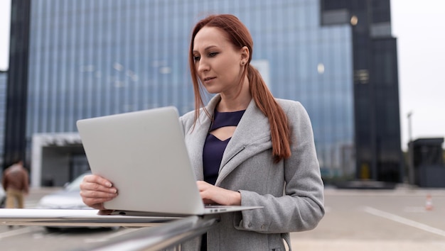 Femme mature aux cheveux roux d'affaires travaillant sur un ordinateur portable en ligne à l'extérieur du bureau sur le fond d'un