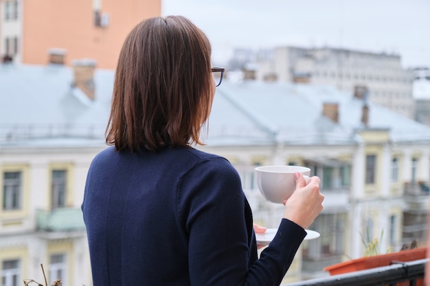 Femme mature debout sur un balcon ouvert avec une tasse de thé, femme regarde la ville en isolement, mis en quarantaine pendant l'infection virale, copiez l'espace