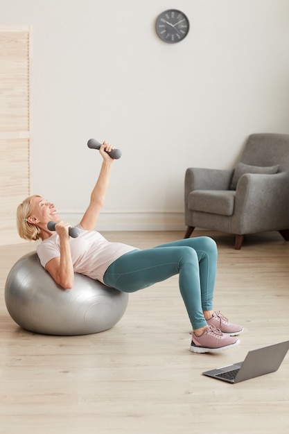 Femme mature exerçant sur le ballon avec des haltères pendant la formation en ligne à la maison