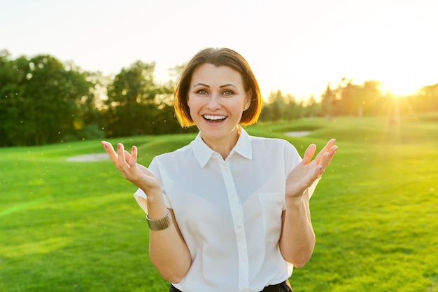 Femme mature parlant positive regardant fond de pelouse verte coucher de soleil du parc de la caméra