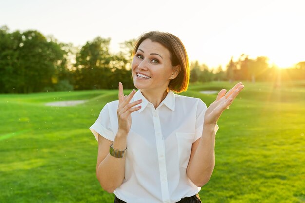 Femme mature parlant positive regardant fond de pelouse verte coucher de soleil du parc de la caméra