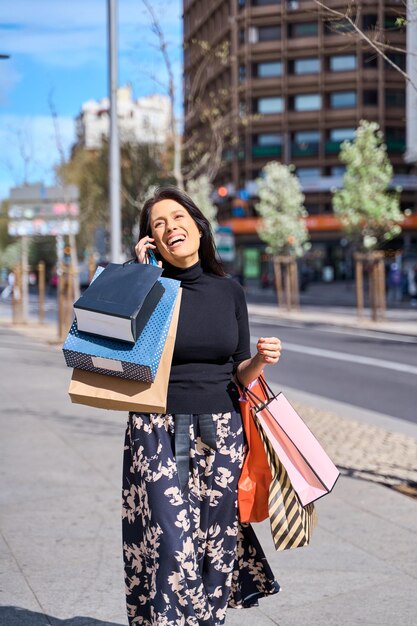 une femme mature parlant sur un téléphone intelligent et riant tout en tenant des sacs d'achat dans ses mains