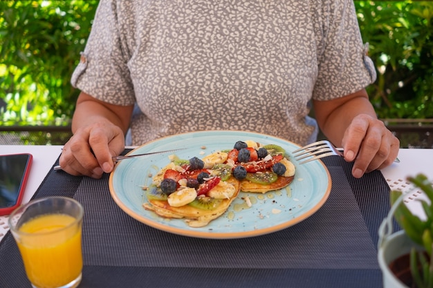 Femme mature avec petit-déjeuner d'été sain, crêpes américaines classiques à la banane, kiwi, baies fraîches et miel