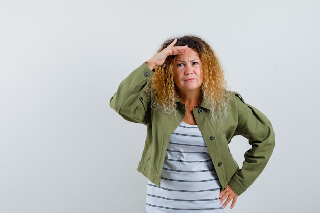 Femme mature à la recherche de loin avec la main sur la tête en veste verte, t-shirt et à la vue de face, focalisée.