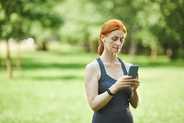 Femme mature rousse dans des écouteurs sans fil debout dans le parc et en choisissant la musique pour la remise en forme à l'aide de smartphone