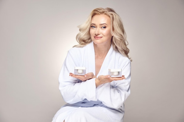 Femme mature satisfaite en peignoir de spa tenant des pots de crème hydratante pour les soins de la peau sur beige isolé