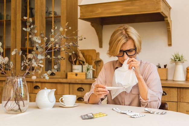 Femme mature senior avec des pilules mesurant la température corporelle avec un thermomètre médical dans la cuisine à la maison. Rhume et grippe, concept de coronavirus covid-19.