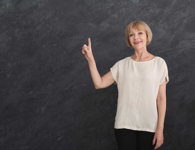 Femme mature souriante pointant vers le haut avec le doigt sur fond gris