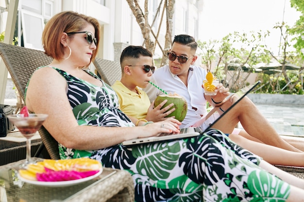 Femme mature travaillant sur un ordinateur portable lorsque son mari et son fils se détendent sur des chaises longues et boivent des cocktails