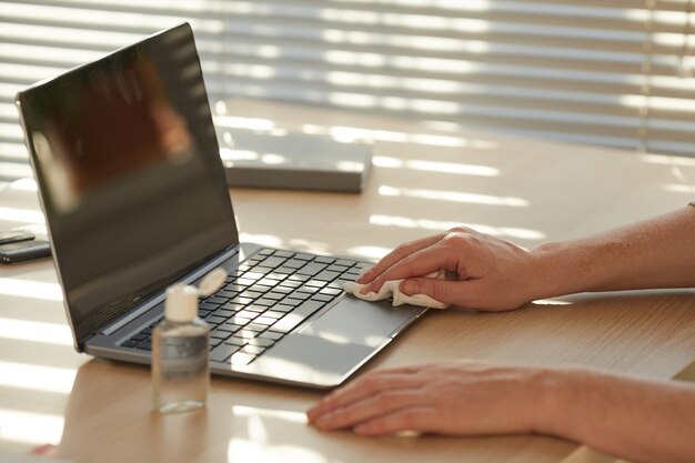 Femme méconnaissable désinfectant un ordinateur portable tout en travaillant au bureau éclairé par la lumière du soleil