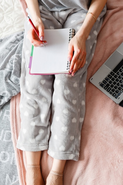 Une femme méconnaissable prend des notes dans le bloc-notes, un ordinateur portable à proximité.