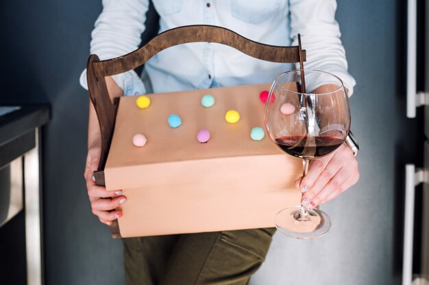 Femme méconnaissable tenant des cadeaux dans un panier en bois et un verre de vin rouge sur fond sombre. Coffret cadeau, concept de fête.