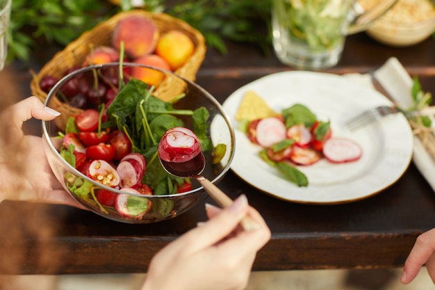 Femme méconnaissable tenant une salade de légumes frais tout en appréciant le dîner avec des amis et la famille à l'extérieur