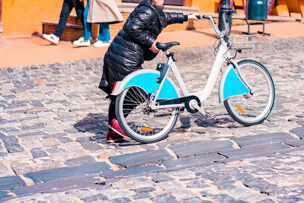 Photo femme méconnaissable en veste noire poussant un vélo de partage sur une large colline pavée dans le centre-ville historique. dur. difficile. pavé. urbain. en haut. rue. partage vélo. vélo. ville vallonnée