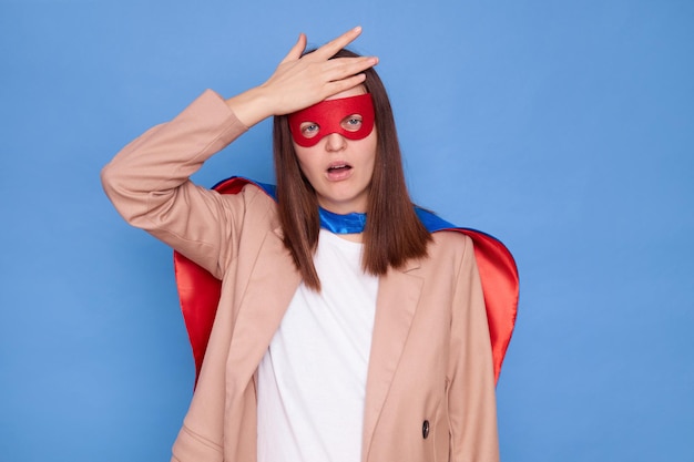 Une femme mécontente et déçue portant un costume de super-héros se tient isolée sur un fond bleu ayant du mal à montrer un geste de paume faciale oubliant quelque chose d'agréable