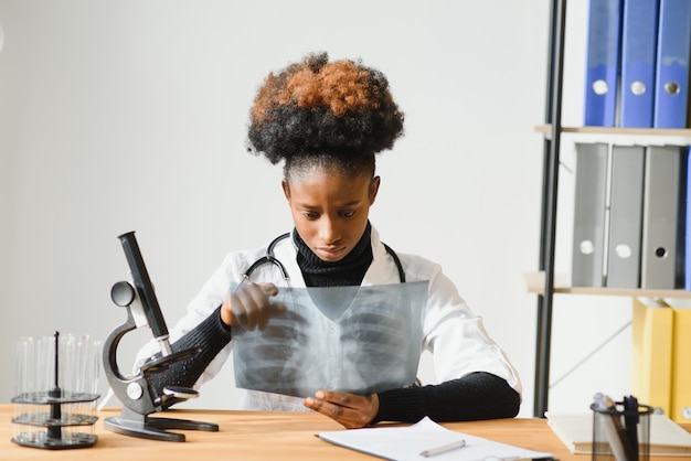 Femme médecin afro-américain portant blouse blanche avec stéthoscope assis derrière un bureau dans le bureau et à la recherche d'image aux rayons x