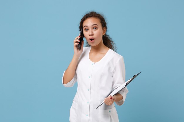 Une femme médecin afro-américaine choquée en robe blanche tient un document médical, parlant sur un téléphone portable isolé sur fond bleu. Concept de santé de la médecine du personnel de santé. Maquette de l'espace de copie.