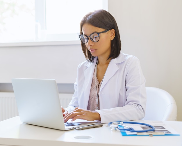 Une femme médecin afro-américaine ciblée à lunettes utilisant un ordinateur portable alors qu'elle était assise sur le lieu de travail dans une clinique médicale, jeune femme médecin afro vêtue d'une blouse blanche travaillant sur ordinateur à l'hôpital
