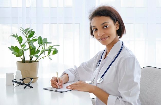 Femme médecin afro-américaine souriante assise à la table du cabinet médical et travaillant sur ordinateur, télémédecine, consultation en ligne des patients