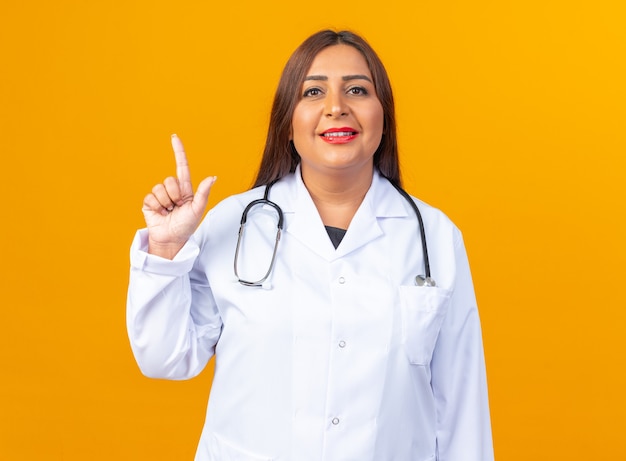 Femme médecin d'âge moyen en blouse blanche avec stéthoscope avec sourire sur un visage intelligent montrant l'index debout sur le mur orange