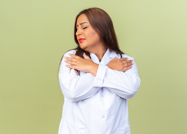 Femme médecin d'âge moyen en blouse blanche avec stéthoscope tenant les mains sur sa poitrine avec les yeux fermés ressentant des émotions positives debout sur un mur vert