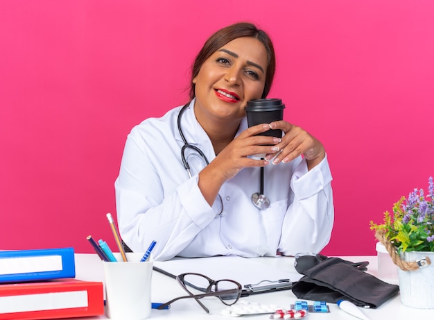 Femme médecin d'âge moyen en blouse blanche avec stéthoscope tenant une tasse de café souriant joyeusement assis à la table avec des dossiers de bureau sur un mur rose