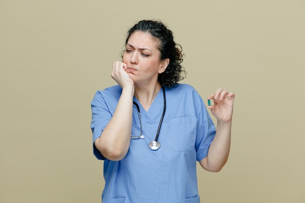 femme médecin d'âge moyen douloureuse portant l'uniforme et le stéthoscope autour du cou montrant la capsule gardant la main sur le menton regardant le côté ayant mal aux dents isolé sur fond olive