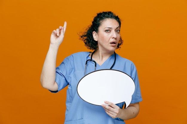 Femme médecin d'âge moyen impressionnée portant un uniforme et un stéthoscope autour du cou montrant une bulle de chat regardant la caméra pointant vers le haut isolée sur fond orange