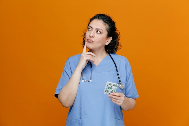 Femme médecin d'âge moyen réfléchie portant un uniforme et un stéthoscope autour du cou tenant un paquet de pilules en gardant le doigt sur le menton en regardant le côté isolé sur fond orange