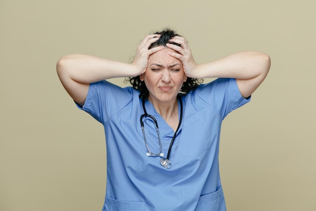 femme médecin d'âge moyen stressée portant un uniforme et un stéthoscope autour du cou en gardant les mains sur la tête avec les yeux fermés isolé sur fond olive