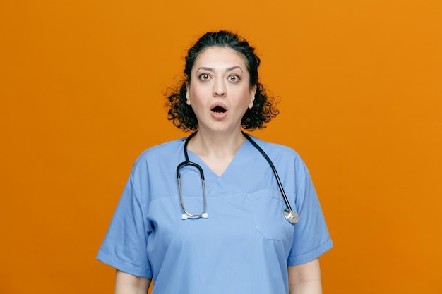 Femme médecin d'âge moyen surprise portant un uniforme et un stéthoscope autour du cou regardant la caméra isolée sur fond orange