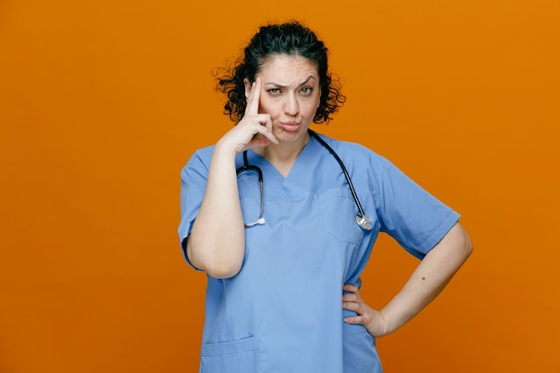 Femme médecin d'âge moyen suspecte portant un uniforme et un stéthoscope autour du cou regardant la caméra en faisant un geste de réflexion tout en gardant la main sur la taille isolée sur fond orange