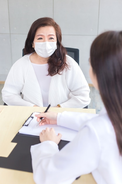 Une femme médecin asiatique enregistre le symptôme du patient sur le document tandis que le diagnostic porte tous deux un masque facial