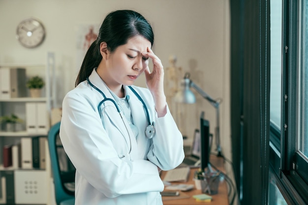 Femme médecin asiatique fatiguée debout à côté d'une grande fenêtre dans le bureau de la clinique. jeune fille épuisée, stagiaire, personnel médical, porter, blanc, manteau, hôpital, lieu travail, confus, pensée, pensif, main, front