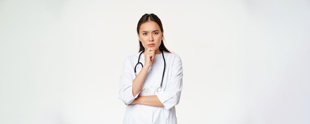 Femme médecin asiatique en uniforme pensant avoir l'air sérieux et inquiet de toucher le menton tout en réfléchissant à l'écoute du patient à l'hôpital fond blanc
