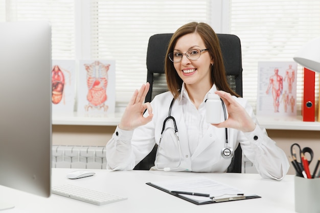 Femme médecin assise au bureau, tenant une bouteille avec des pilules blanches, travaillant avec des documents médicaux dans un bureau léger à l'hôpital