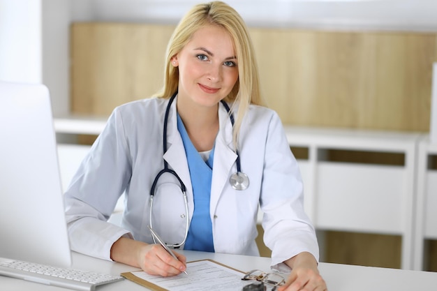 Femme médecin au travail assise au bureau à l'hôpital ou à la clinique Médecin gai blond remplissant le formulaire de dossier médical Données et meilleur service en médecine et en soins de santé