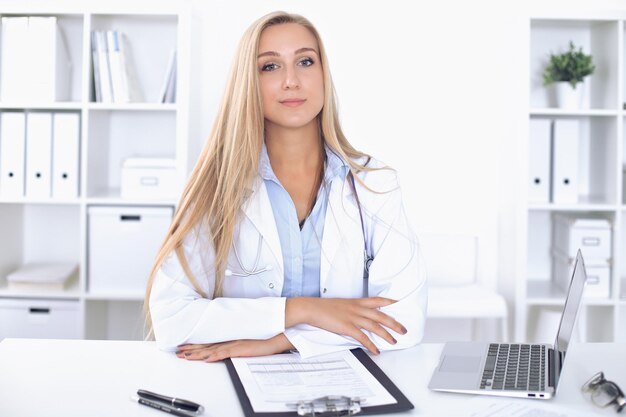 Femme médecin blonde assise à la table à l'hôpital et regardant la caméra.