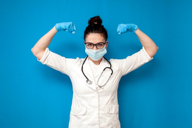 Femme médecin en blouse médicale et masque médical montre un geste de force sur fond bleu