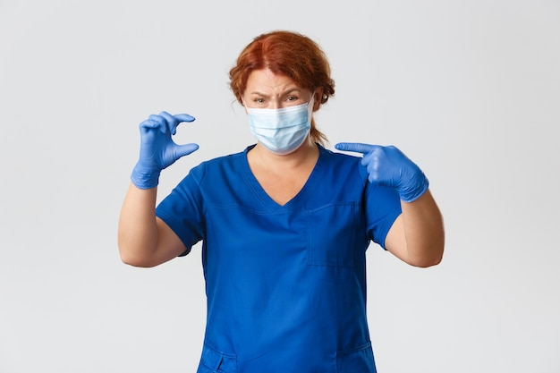 Femme médecin bouleversée et déçue, infirmière d'âge moyen en masque facial, gants en caoutchouc pointant vers quelque chose de petit ou petit avec une expression triste.