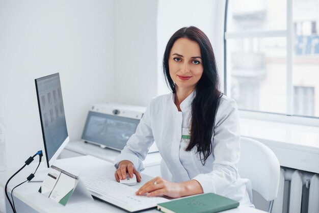 Une femme médecin brune est assise dans un bureau moderne par ordinateur et regarde la caméra.