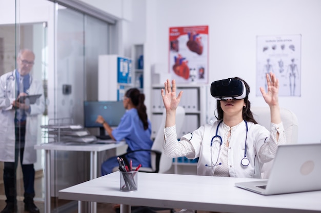 Femme médecin caucasienne en robe blanche et casque VR assis au bureau dans l'armoire en glissant et en faisant défiler les mains en l'air. Concept de réalité virtuelle. Santé futuriste. Docteur travaillant à l'avenir.