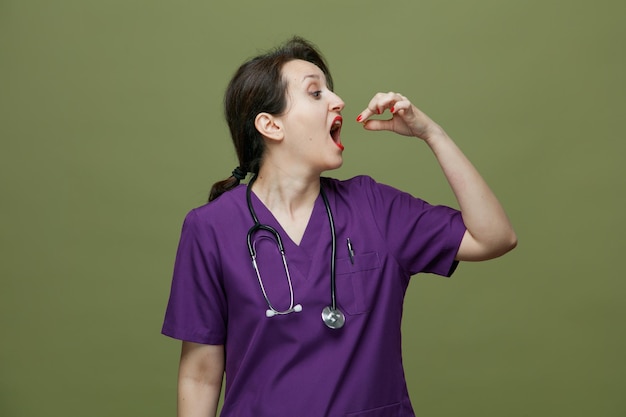 Femme médecin concentrée d'âge moyen portant un uniforme et un stéthoscope autour du cou tournant la tête à l'autre en prenant la pilule isolée sur fond vert olive