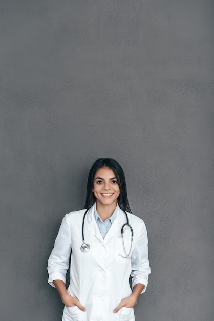 Femme médecin confiante. Jeune femme médecin confiante en blouse blanche tenant les mains dans les poches et souriant en se tenant debout sur fond gris