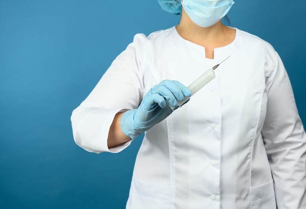 Une femme médecin dans une blouse blanche et un masque se dresse sur un mur bleu et tient une seringue en plastique, conception de la vaccination